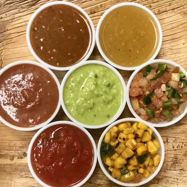 Seven small white bowls of various Mexican salsas and toppings arranged on a wooden table.